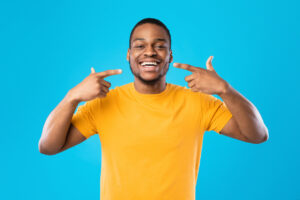 The image shows a Black man pointing cheerfully at his smile to represent how to get the most out of Invisalign treatment.