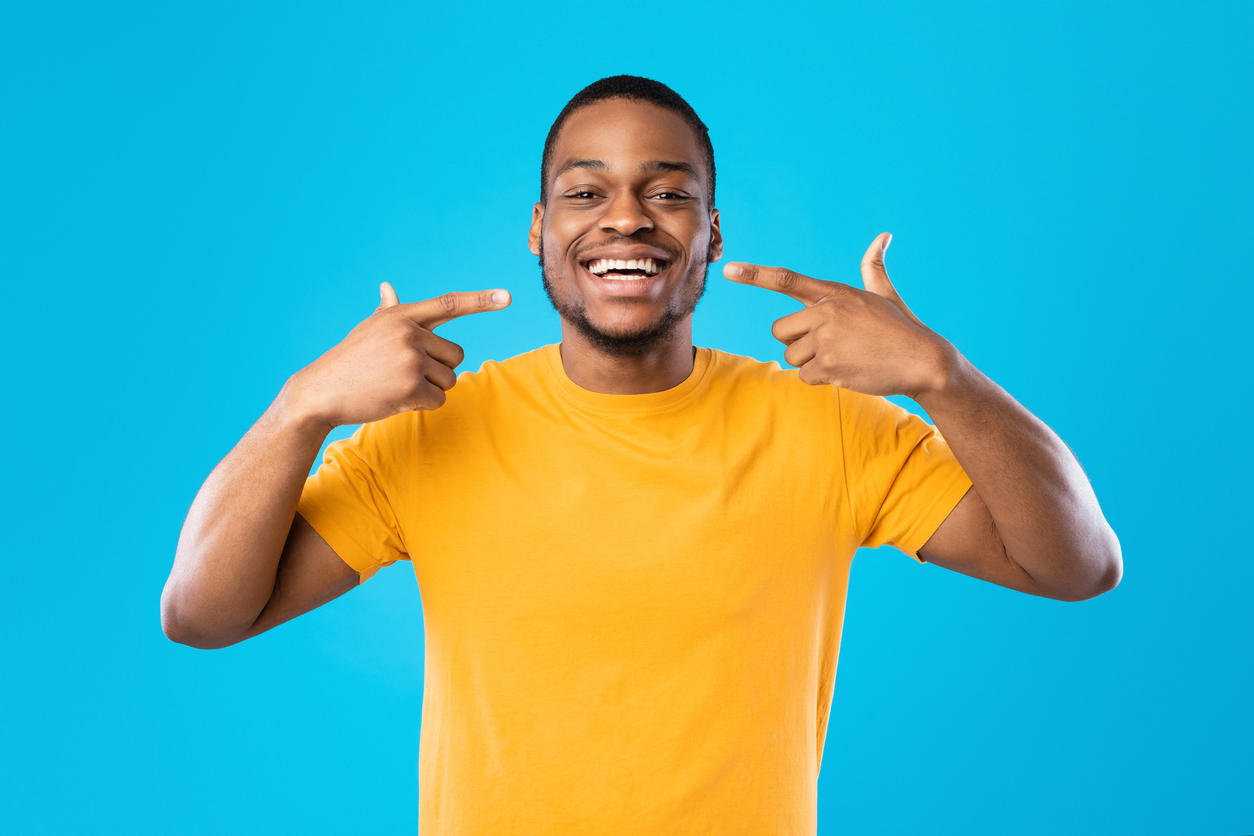 The image shows a Black man pointing cheerfully at his smile to represent how to get the most out of Invisalign treatment.